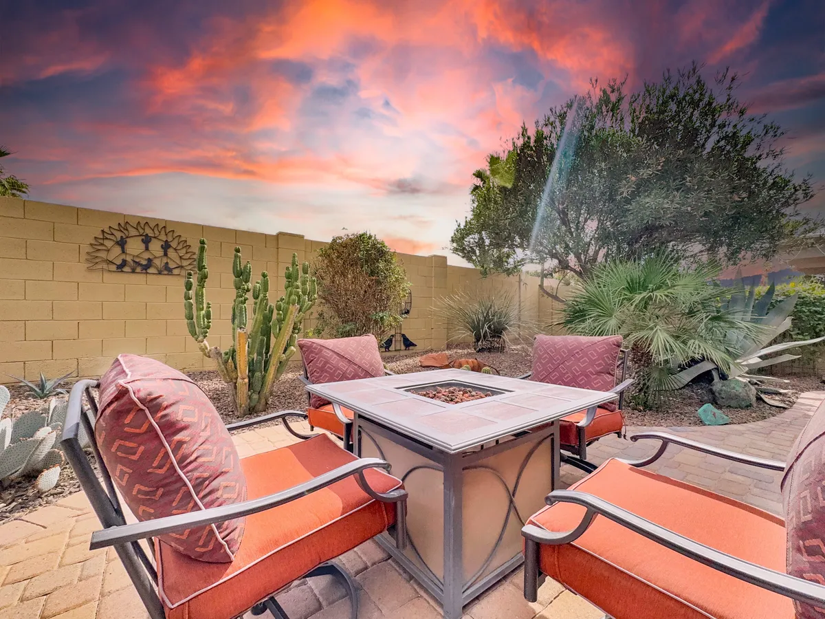 The scene centers on a cozy outdoor fire pit with surrounding seating, framed by native cacti and set beneath a vibrant sunset sky. The atmosphere is both relaxed and social, ideal for gatherings or intimate conversations under the stars.