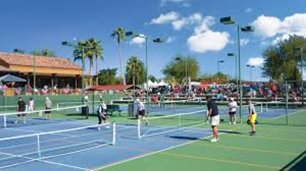 This image features lively tennis courts surrounded by palm trees, with active players and engaged spectators. The nearby clubhouse hints at additional recreation and social opportunities within the community, all framed by clear desert skies.