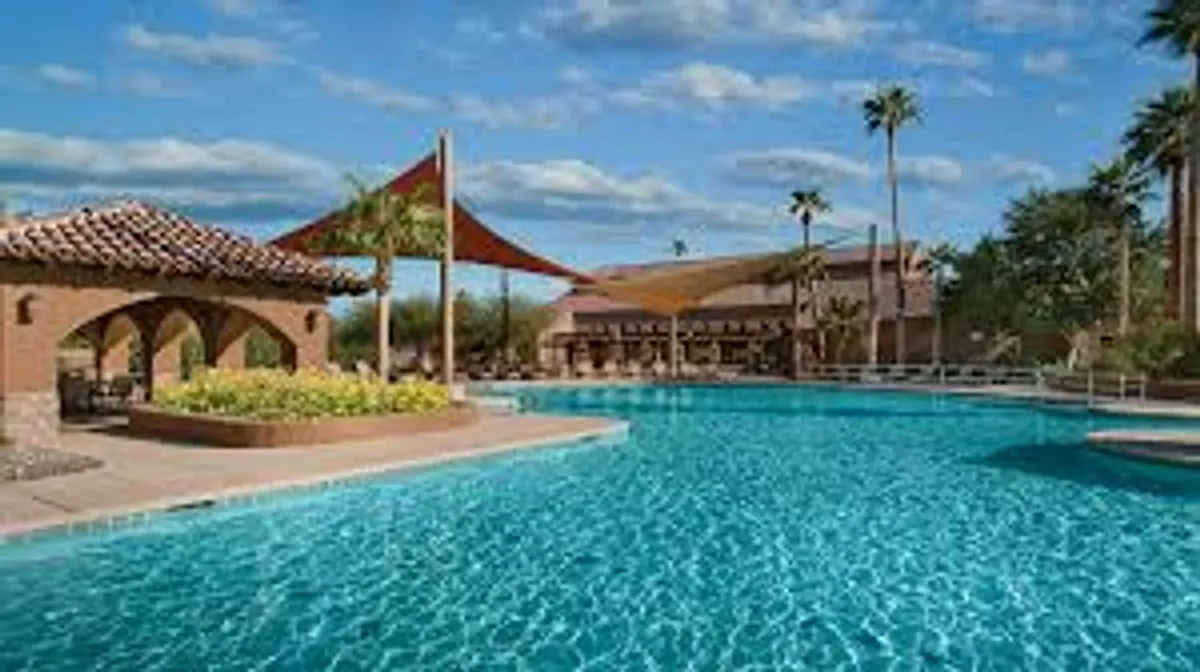 A sunlit, resort-style swimming pool surrounded by lush landscaping and a graceful shade structure sets the scene. The poolside area is perfect for relaxation, sunbathing, or gathering with friends, evoking the resort vibe described in the text.