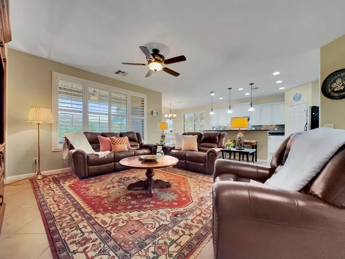 This image features a spacious living room with leather sofas arranged around a round coffee table and an ornate area rug. Large windows let in abundant natural light and showcase tranquil garden views outside. Pendant lights add a warm ambiance, and the room is clearly designed for both social gatherings and quiet relaxation.