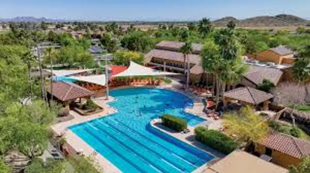 A vibrant community swimming pool area complete with lounge chairs, shade structures, and mature palm trees, evoking a resort-like atmosphere. The area is set for relaxation, swimming, and socializing, highlighting the breadth of amenities available at Robson Ranch.