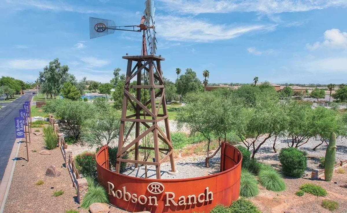 The iconic Robson Ranch entrance is marked by a windmill, landscaped greenery, and a welcoming sign. Walkways and natural desert elements invite exploration and signal a distinct sense of place and community pride.